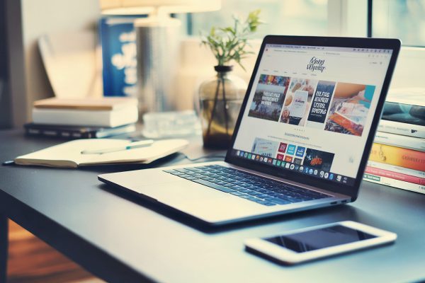 Moderner Arbeitsplatz mit silbernem Laptop, der eine Website zeigt, Smartphone, Bücherstapel und kleiner Pflanze im Glasgefäß bei natürlichem Licht