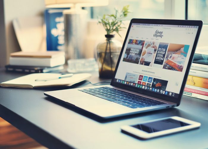 Moderner Arbeitsplatz mit silbernem Laptop, der eine Website zeigt, Smartphone, Bücherstapel und kleiner Pflanze im Glasgefäß bei natürlichem Licht